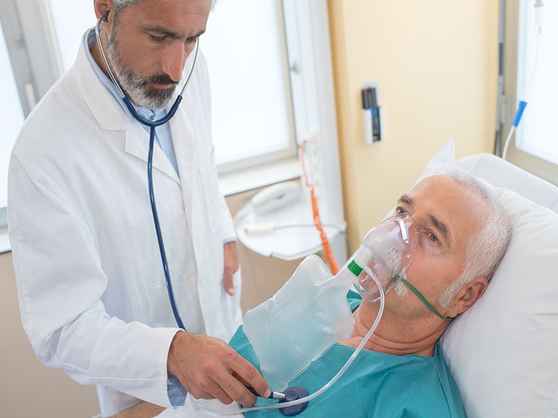 man in hospital bed with breathing mask getting his heart listened to by doctor