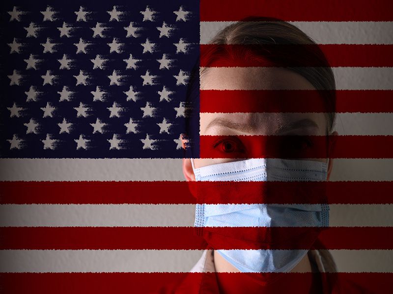 female doctor wearing blue medical mask standing behind an american flag