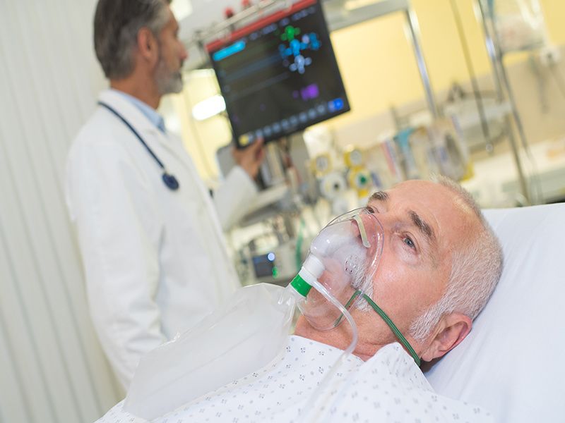 older man in hospital bed with breathing mask while male doctor checks vitals on monitor screen
